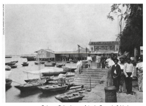 Fotografia antiga em tons de sépia de um porto com barcos pequenos, escadaria e pessoas à beira da água.