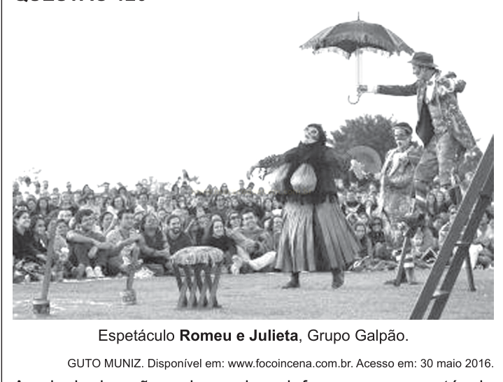 Fotografia em preto e branco de atores em cena ao ar livre diante de uma plateia, com destaque para um ator de cartola com guarda-chuva sobre uma escada.