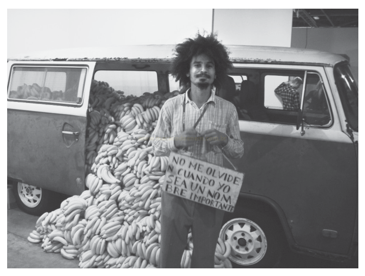 Fotografia em preto e branco de um homem segurando um cartaz diante de uma van carregada de bananas.