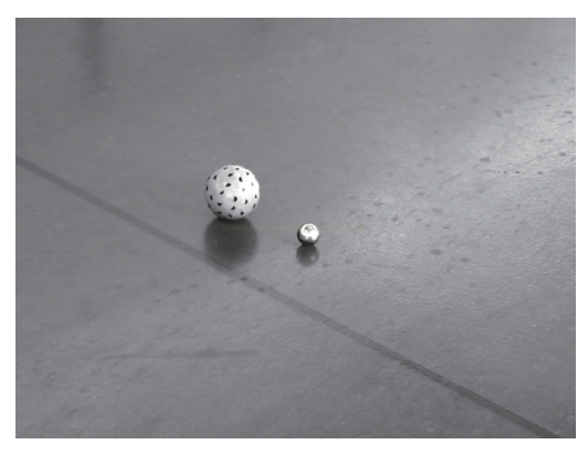 Fotografia em preto e branco de duas bolas brancas com pontos pretos, uma grande e outra muito pequena, sobre o piso.