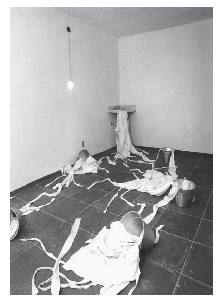 Fotografia em preto e branco de uma sala com tiras de tecido espalhadas pelo chão e baldes metálicos.