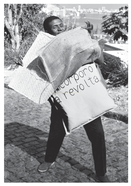 Fotografia em preto e branco de um menino carregando duas grandes almofadas, uma com os dizeres "o corpo a revolta".