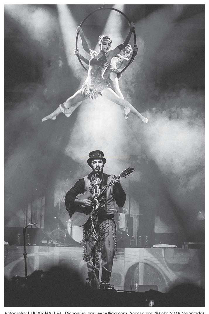 Fotografia em preto e branco de número de circo: acrobata em aro suspenso na altura do teto e músico de cartola tocando violão no palco.