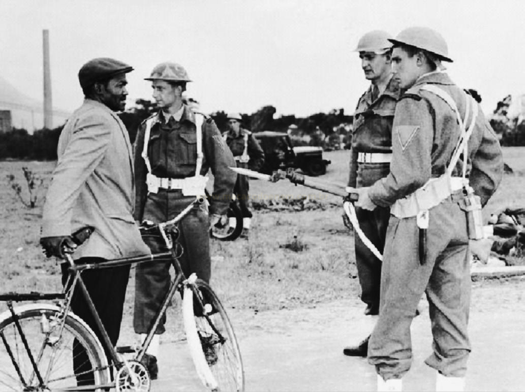 Fotografia em preto e branco de um homem com bicicleta diante de militares, um deles apontando um rifle.