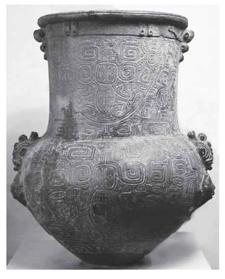 Fotografia em preto e branco de vaso antigo de metal com relevos geométricos e alças laterais.