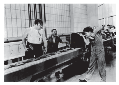 Fotografia em preto e branco de três trabalhadores em uma bancada de oficina, um deles erguendo uma ferramenta.