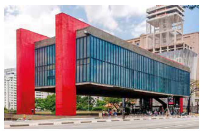 Fotografia de um edifício moderno com bloco de vidro suspenso sobre pilares vermelhos, em uma avenida.