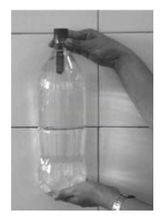 Fotografia em tons de cinza de mãos segurando uma garrafa plástica transparente diante de uma parede de azulejos.