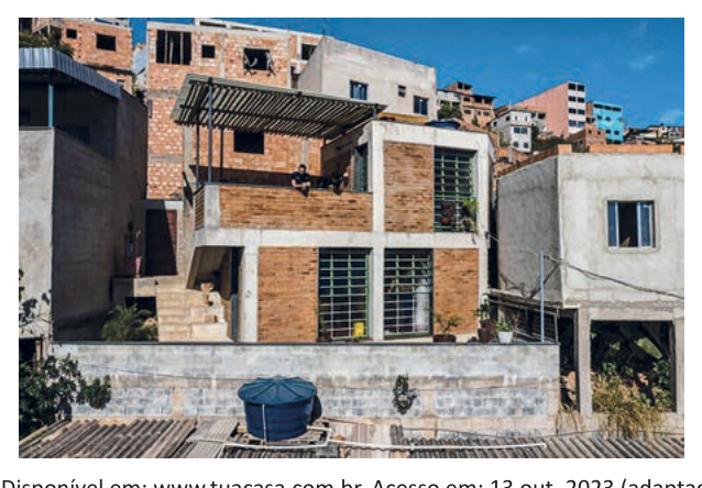 Fotografia colorida de uma casa de tijolo aparente em encosta, com varanda e outras construções ao redor.