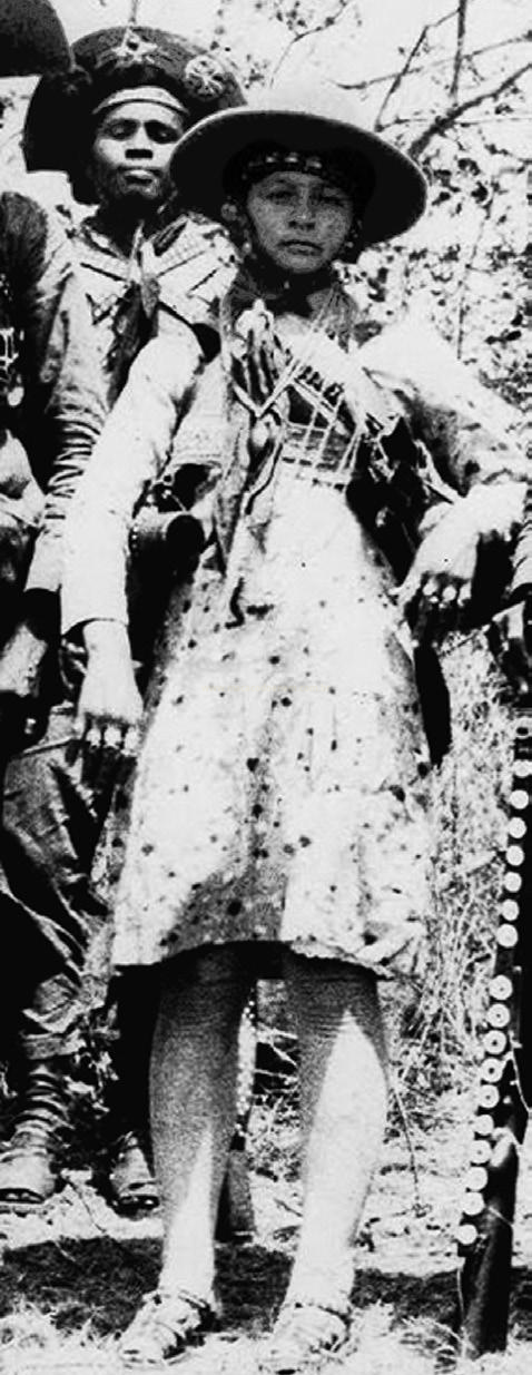 Fotografia antiga em preto e branco de uma pessoa de chapéu de aba larga e túnica clara, com outra pessoa fardada ao fundo.