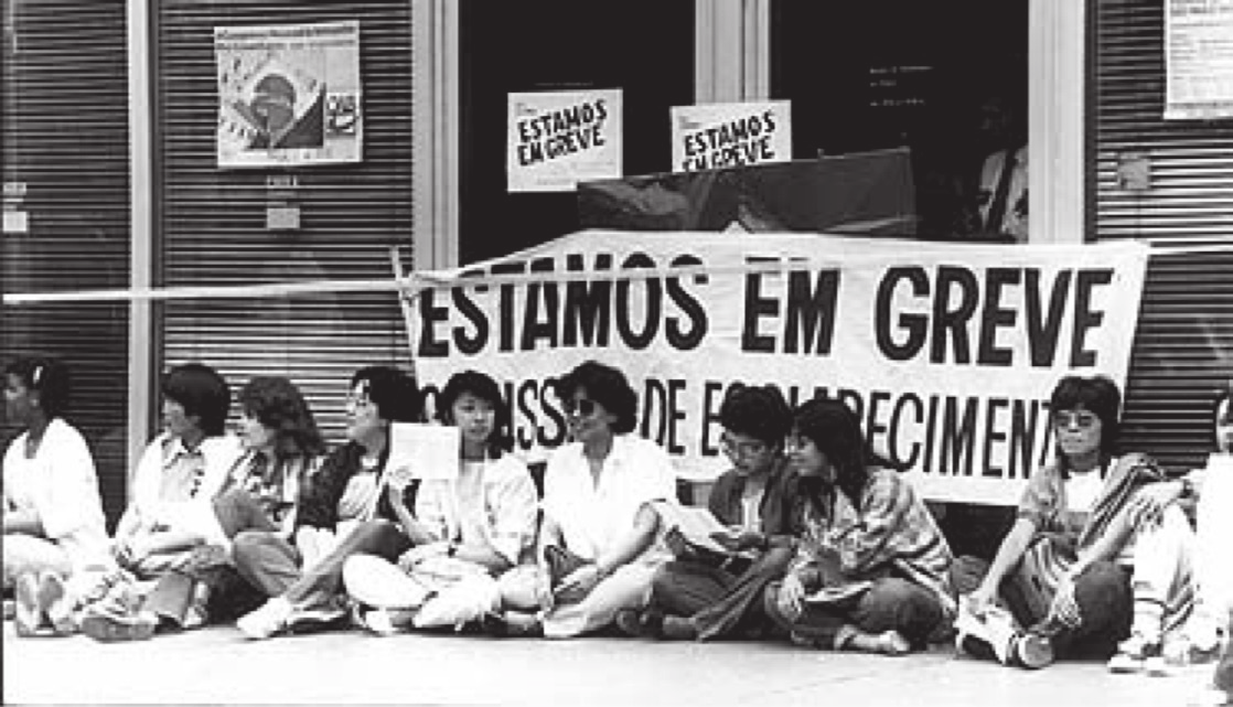 Fotografia em preto e branco de mulheres sentadas no chão diante de um prédio, atrás de uma faixa "Estamos em greve".