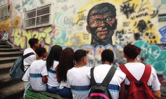 Fotografia de estudantes de uniforme, vistos de costas, sentados diante de um mural grafitado com o retrato de um homem negro.